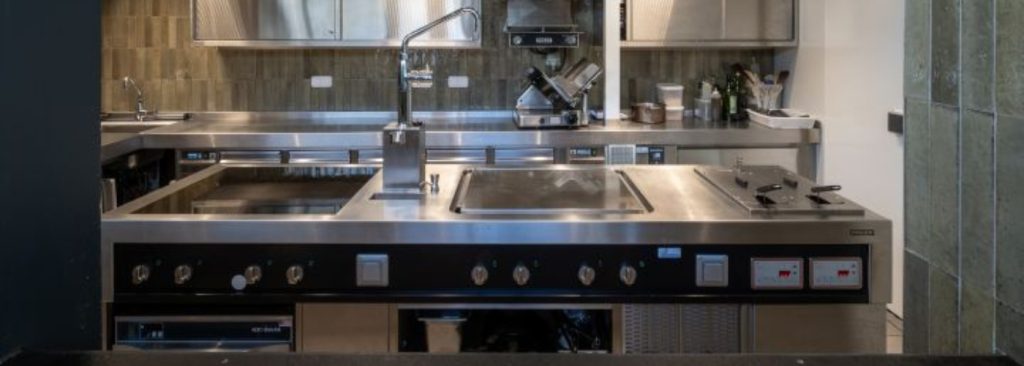 Omslagfoto van Professionele keuken van Den Heerd met centraal Palux-eiland, inox werkbladen en hangende kookpotten.